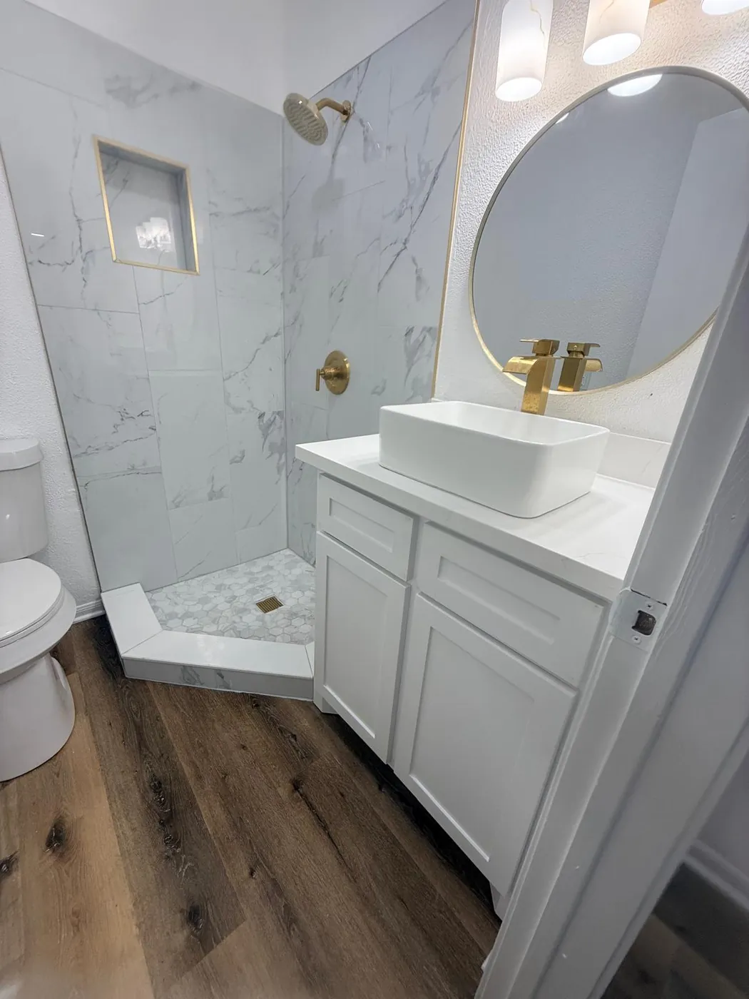 Marble-look shower with gold fixtures, vessel sink and a round mirror.